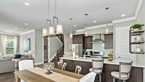 A modern kitchen with dark cabinetry, a central island, pendant lighting, and a dining area with a wooden table and chairs. - Golf Cart ~ Private Pool ~ Near Beach ~ Grill (Santa Rosa Beach)