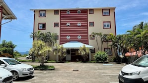 A multi-story building with a central entrance, parking lot, and surrounding palm trees. - T2 40 m2 Anse Mitan - Panorama bay of Fort-de-France (Les Trois-Îlets)
