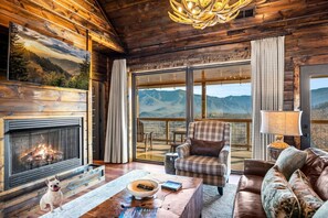 A log cabin interior with a fireplace, a dog, a plaid armchair, and a view of mountains through large windows. - Camp: Dog-Friendly•MtnView•HotTub•Luxury•2KingStes (Gatlinburg)