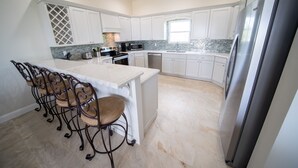 Una cocina moderna con armarios blancos, una isla central con sillas, un refrigerador de acero inoxidable y un ventanal que proporciona luz natural.