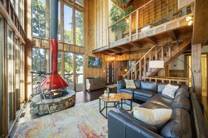 Un vaste espace de vie agrémenté d’une cheminée en pierre, d’un canapé en cuir et d’une grande fenêtre donnant sur une forêt.