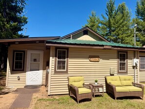 A cabin with a green roof, a welcome sign, a door, two wicker chairs with cushions, and a small table with a plant.