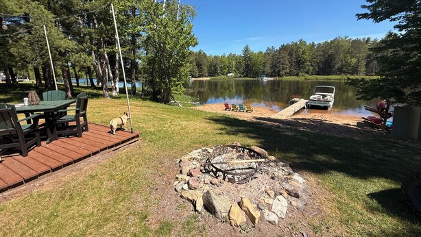 En terrass vid sjön med ett bord och stolar, en eldstad och en brygga med en båt.
