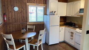 Ett kompakt kök med vita skåp, ett kylskåp, en spis och ett fönster med persienner.