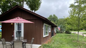 En liten brun byggnad med ett utebord och stolar, en amerikansk flagga och en gräsplätt.