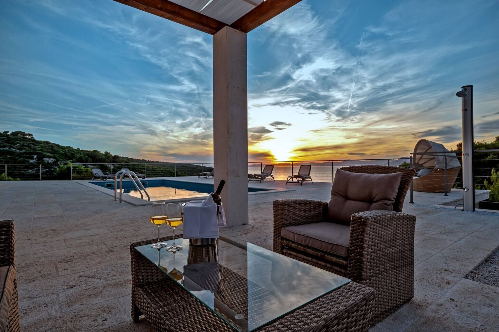 A rooftop patio with a glass table, wicker chairs, a pool, and a sunset view.