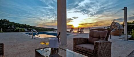 A rooftop patio with a glass table, wicker chairs, a pool, and a sunset view.