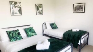 Une chambre double avec deux lits, chacun agrémenté d’un oreiller à motifs de feuilles vertes, de draps blancs et d’un plaid vert foncé. Trois tableaux encadrés ornent le mur.