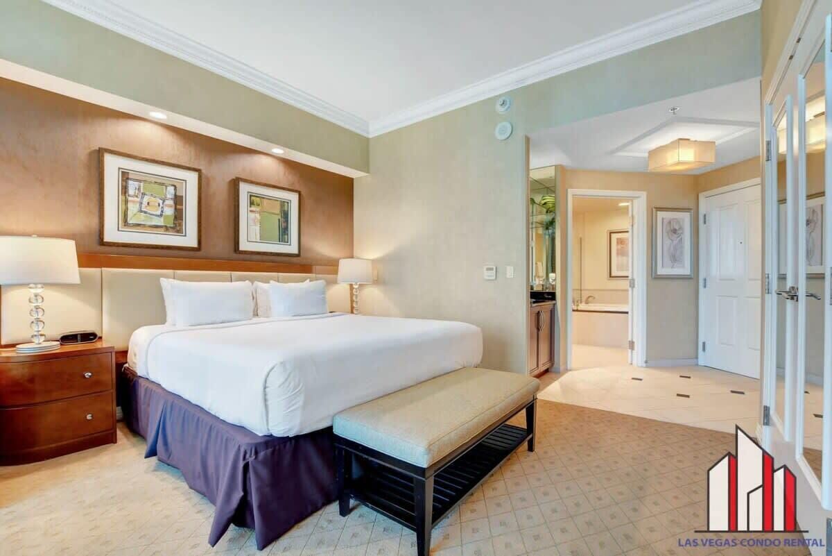 A hotel room with a large bed, a bench, a nightstand, and a bathroom visible through an open door.