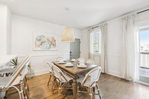 A dining area with a wooden table, wicker chairs, a hanging light fixture, and a large window with curtains.