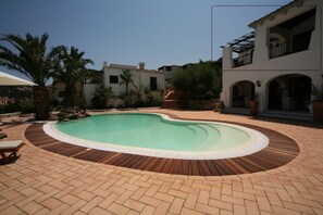 Une piscine dotée d’une terrasse en bois, entourée de palmiers et d’un patio en briques.