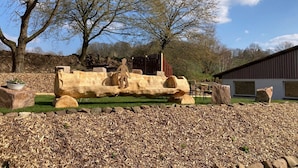 Une sculpture représentant une figure posée sur une bûche, dans un espace aménagé avec des arbres et un bâtiment en arrière-plan.