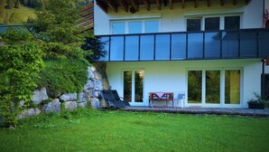 Une maison moderne à deux étages, dotée d’un mur de soutènement en pierre, d’une pelouse et d’une terrasse avec une table et des chaises.