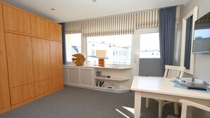 A room with a wooden wardrobe, a white desk with a lamp, a chair, and a window with curtains.