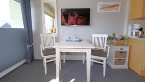 A small, clean room with a white table and two chairs, a flat-screen TV mounted on the wall, and a kitchenette area with a coffee maker and wicker baskets.