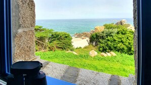 A room with a stone wall and a window overlooking a beach with greenery and the sea.