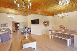 Uma sala de estar ampla com teto de madeira, uma mesa de jantar com bancos, um sofá e uma TV fixada na parede.