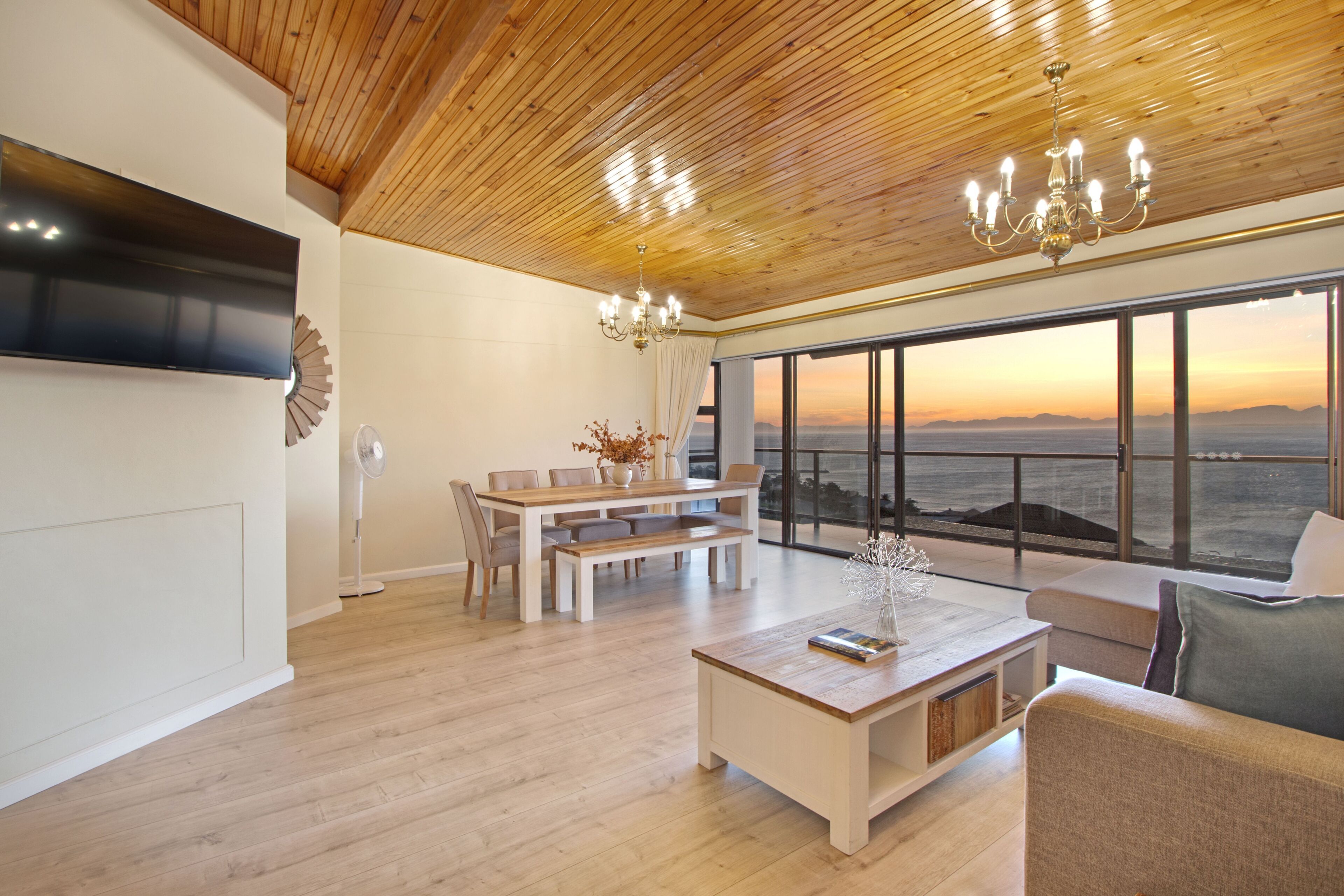 Amplia sala de estar con techo de madera, una gran mesa de comedor, un sofá y vista al océano al atardecer.