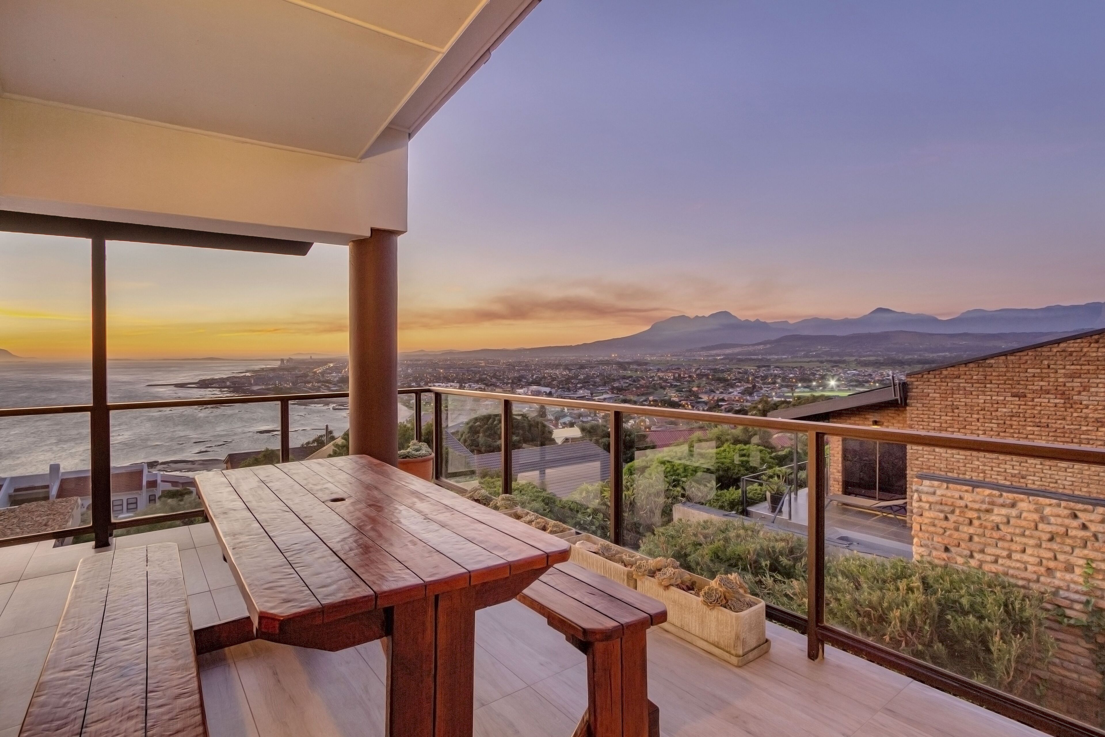 Una terraza de madera con una mesa y bancos, con vistas a un paisaje urbano y montañas durante el atardecer.