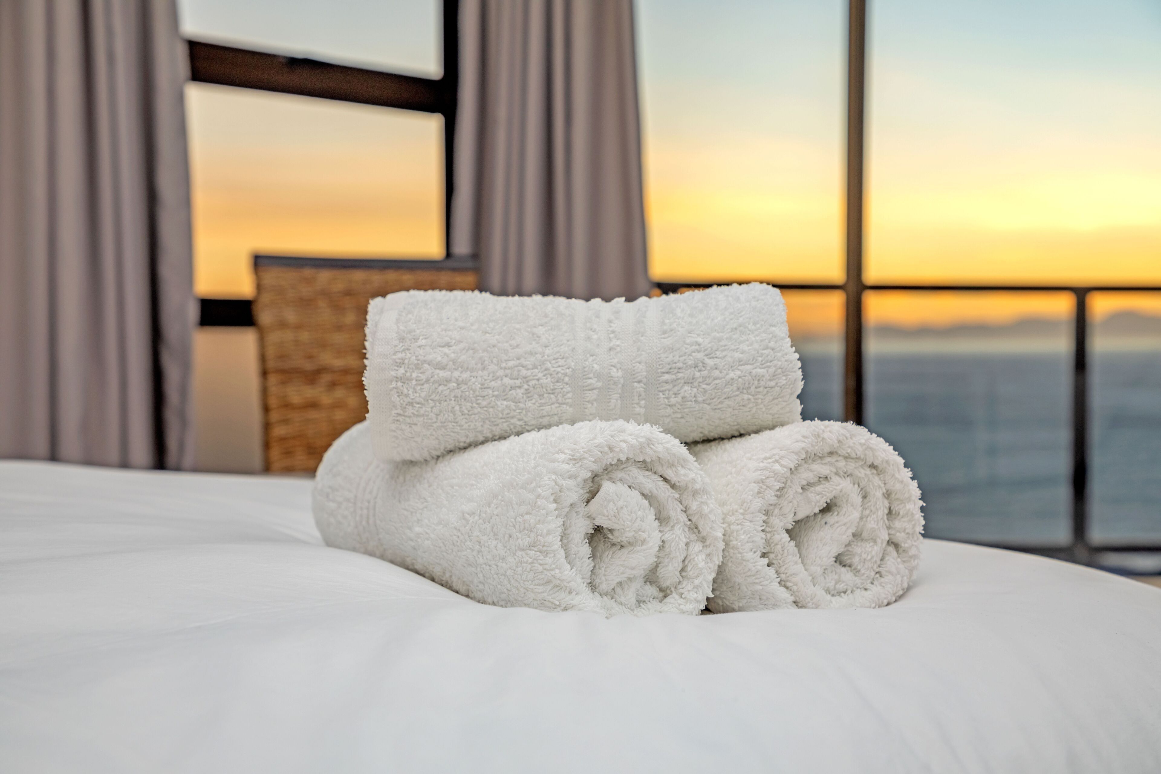 A neatly made bed with white towels and a view of the sunset through large windows.
