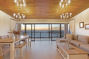 Un vaste salon doté d’un plafond en bois, de grandes fenêtres offrant une vue sur la mer, d’une table à manger avec des bancs et d’un canapé.