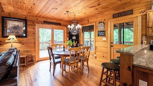 Interior de una cabaña con una mesa de comedor de madera, sillas, una araña, un sofá y una zona de cocina con encimera de granito.