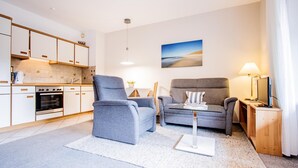 Een moderne woonkamer met twee grijze fauteuils, een glazen salontafel en een bank. Er is een keuken met witte kasten en een ingebouwde oven. Aan de muur hangt een schilderij van een landschap, en op het bijzettafeltje staat een lamp.