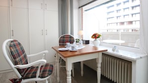 A dining area with a table set for two, a plaid chair, a white chair, a radiator, and a window with a view of a building.