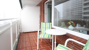 A balcony with a white table and striped chairs, a white refrigerator, and a view of buildings through a window.