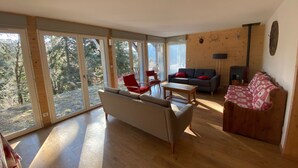 A spacious living room with large windows, a sofa, a red chair, a wooden coffee table, and a fireplace. - Appartement Rocher du Cerf T5 (Le Lioran)