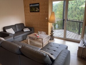 Un salon moderne avec un canapé gris, une table basse sur laquelle sont posés des verres à vin, et un balcon offrant une vue sur une forêt.