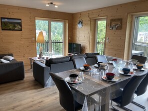 Ein gemütliches Zimmer mit Esstisch für vier Personen, einer Couch, einem Fernseher und großen Fenstern mit Blick ins Grüne.
