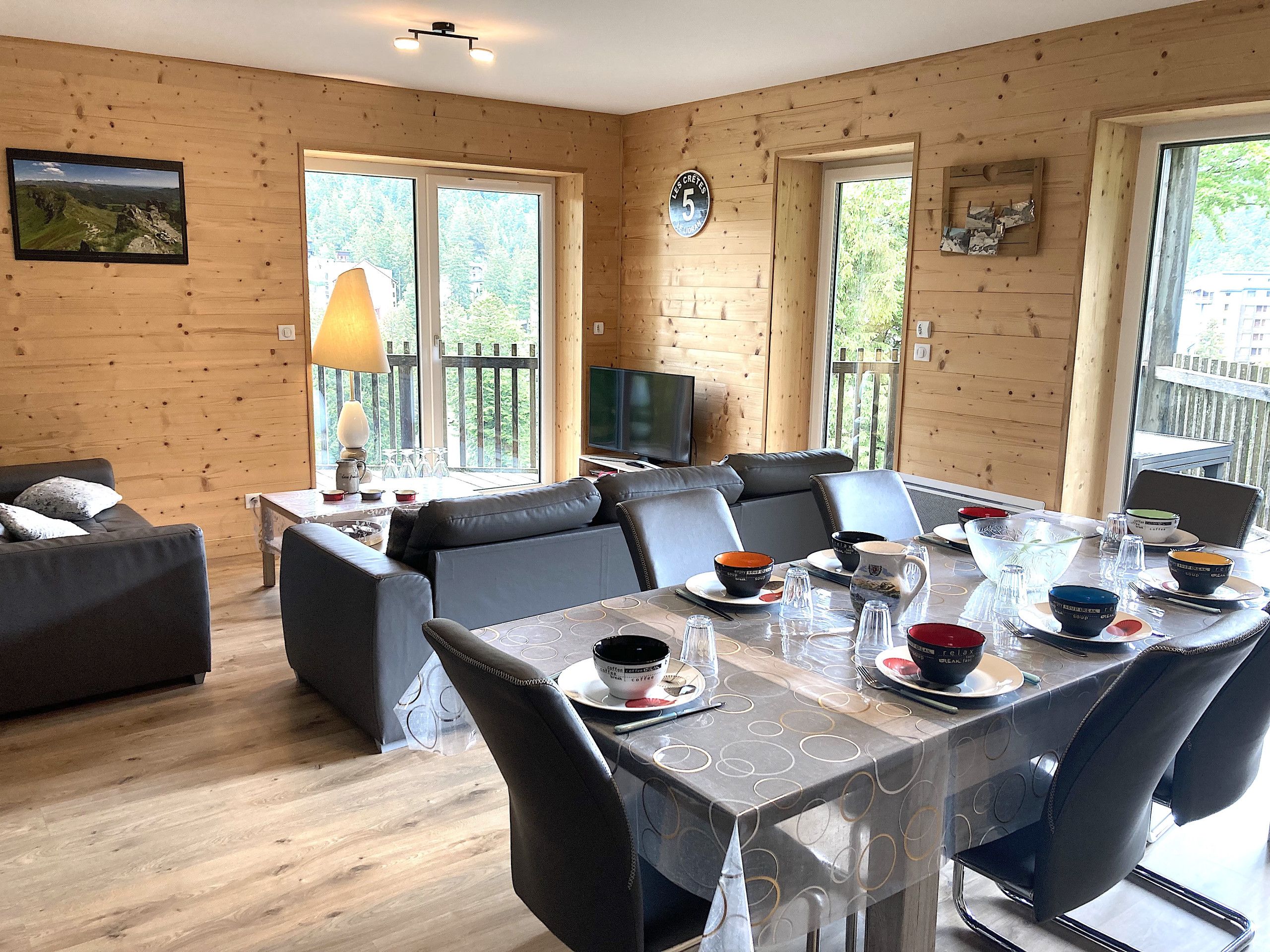 A dining area with a table set for four, a sofa, and a television in a room with wooden walls and large windows.