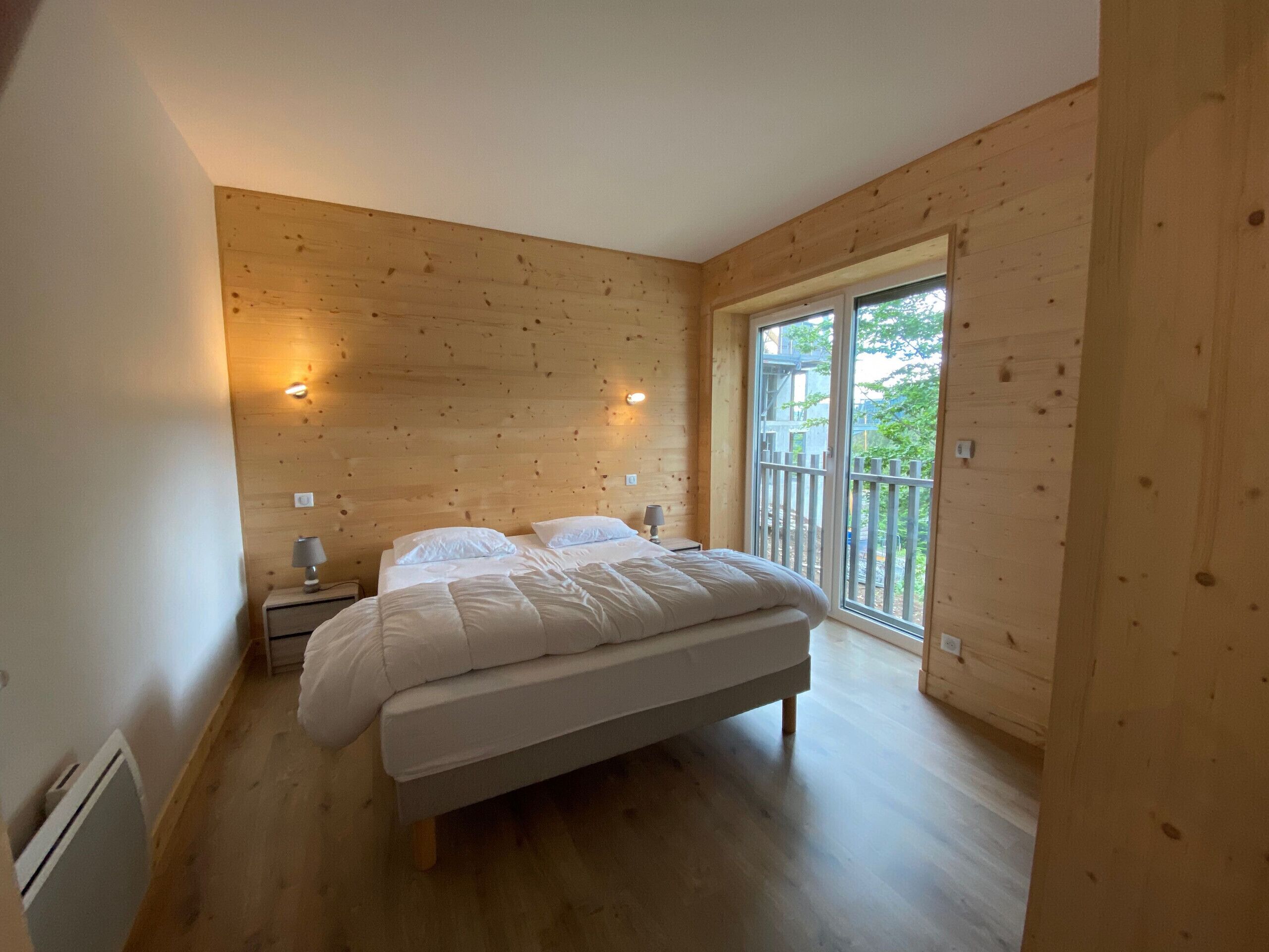 Une chambre avec un plancher en bois, un lit recouvert de linge de lit blanc, deux lampes de chevet et une grande fenêtre donnant sur l’extérieur.