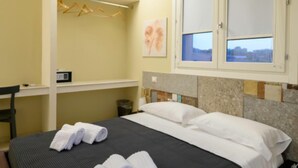 A hotel room with a bed, a desk, a chair, a window, and a wall-mounted radio.