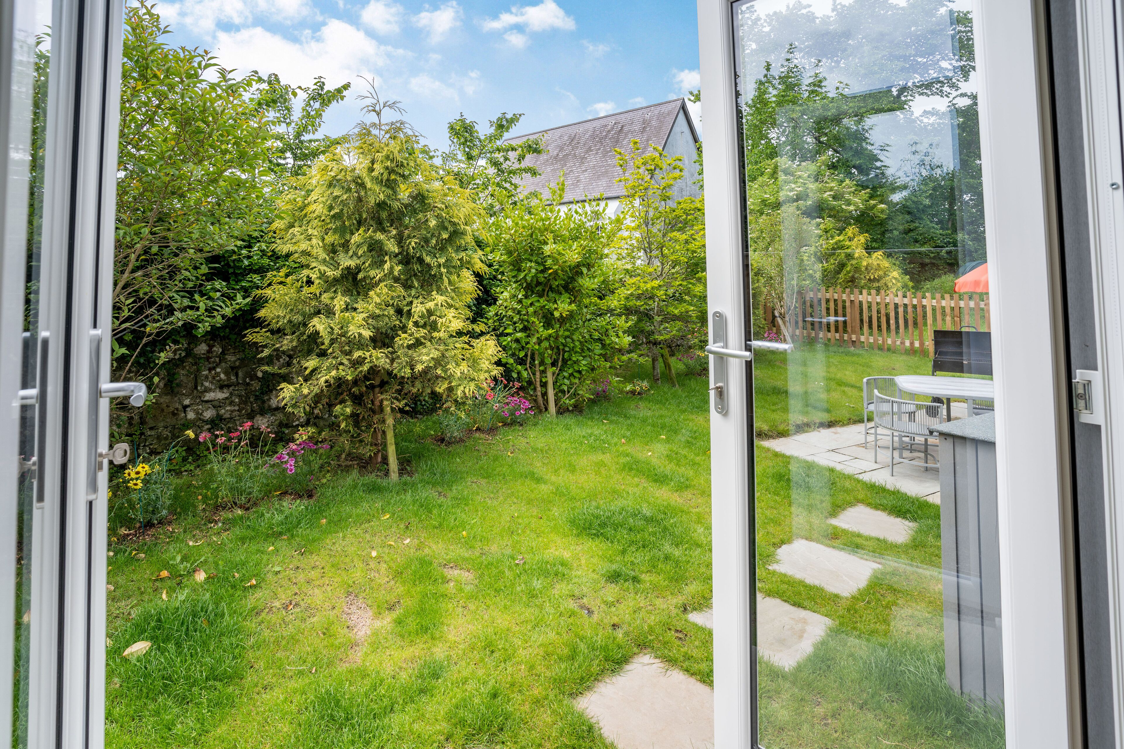A view through a glass door into a backyard with a lawn, stepping stones, a wooden fence, and a table with chairs.