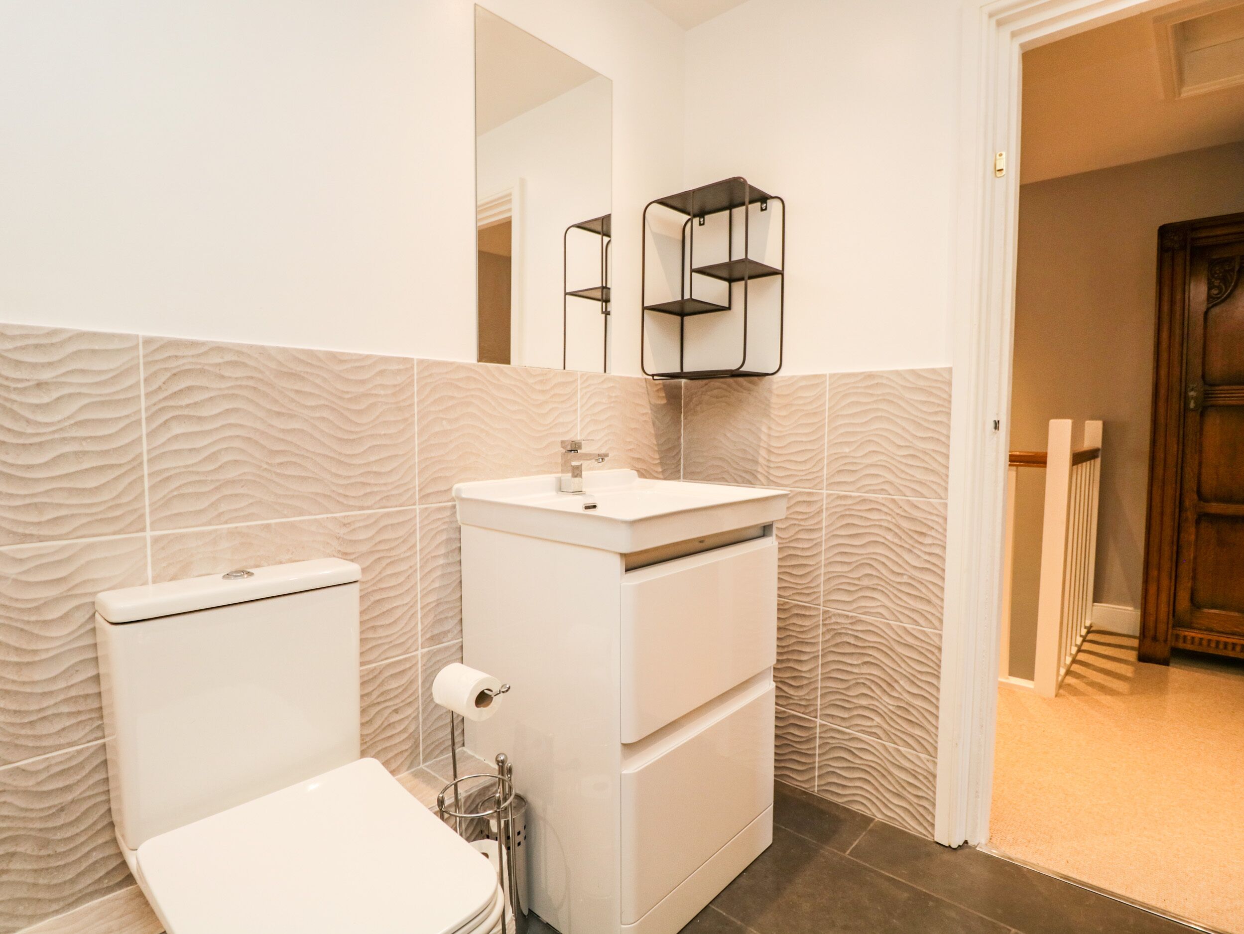 A bathroom with a toilet, sink, and mirror. There is a towel rack and a trash can. The walls have a textured pattern.