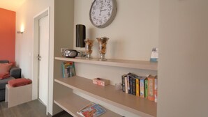 Un pasillo con un reloj, estantes con libros y objetos decorativos, una radio y una puerta que da a otra habitación.