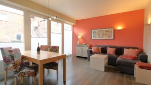 Una sala de estar moderna con una mesa de comedor de madera, sillas coloridas con estampado, un sofá gris con cojines naranjas y una pared de acento roja.