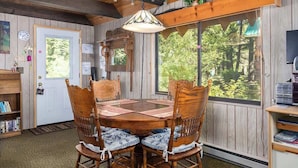 A wooden interior with a round table, chairs, a bookshelf, and a large window with a view of trees.