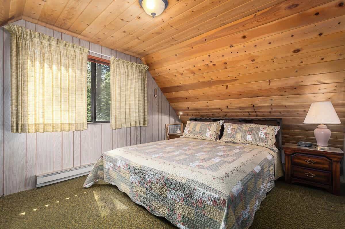 A bedroom with a wooden ceiling, a bed with a floral quilt, a nightstand with a lamp, and a window with curtains.