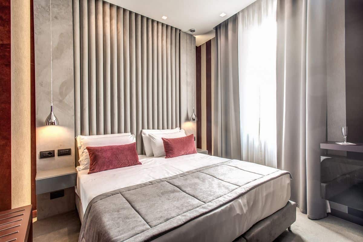 Une chambre d’hôtel moderne avec un grand lit, une table de chevet, une lampe fixée au mur et une vue sur l’extérieur par la fen être.