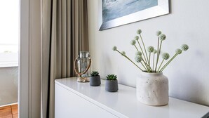 Una consola blanca con un jarrón blanco que contiene plantas verdes, un tarro de vidrio con asa de cuerda y un cuadro en la pared.