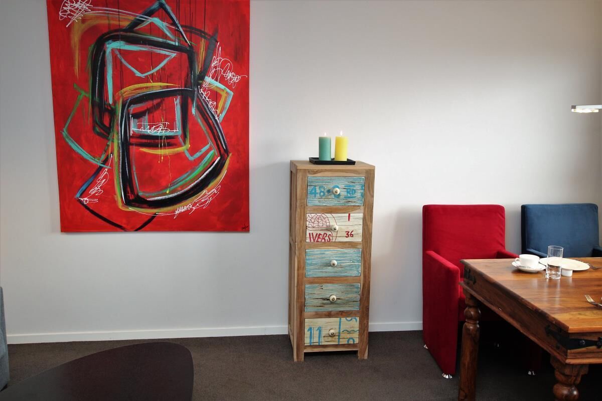 Ein Essbereich mit einem Holztisch, gedeckt für zwei Personen, einem roten und blauen Stuhl, einer Holzkabine mit Schubladen und einem großen abstrakten Gemälde an der Wand.