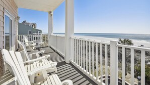 A covered balcony with white railings, a table, and chairs, overlooking a beach with a clear view of the ocean.