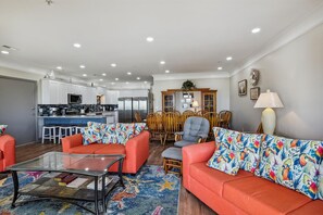 A modern living room with a glass coffee table, two orange sofas with colorful patterned cushions, a dining area with wooden chairs, and a kitchen with white cabinets and stainless steel appliances.