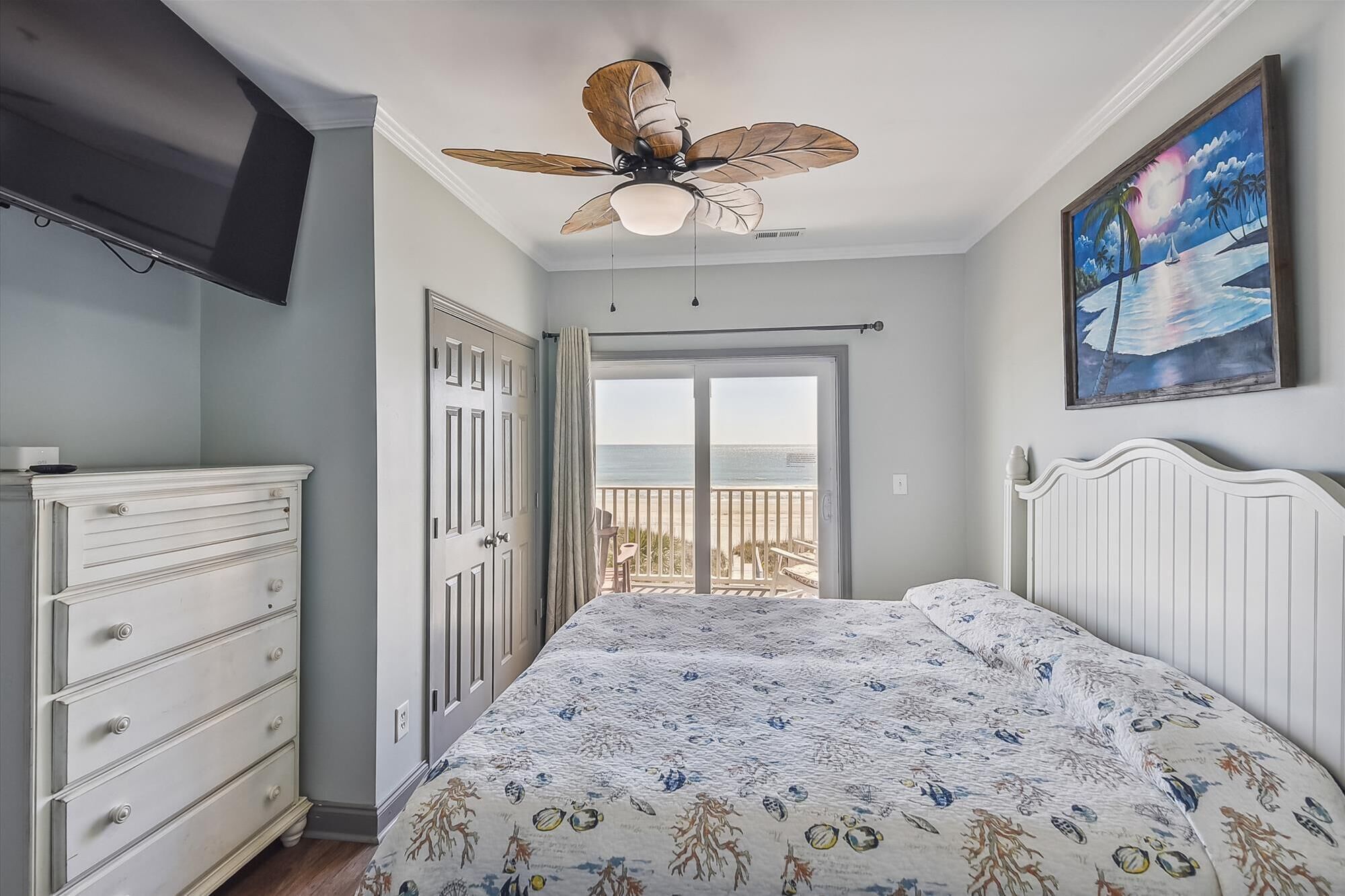A bedroom with a bed, a dresser, a ceiling fan, a painting, and a view of the beach through an open door.