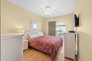 Un dormitorio con cama, mesita de noche, lámpara, ventilador de techo, televisor y ventana con cortinas.