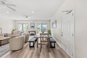 A spacious living room with a ceiling fan, a dining area with a wooden table and benches, and a view of the outdoors through large windows.