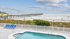A pool area with lounge chairs, a pier extending into the ocean, and a beach in the background.